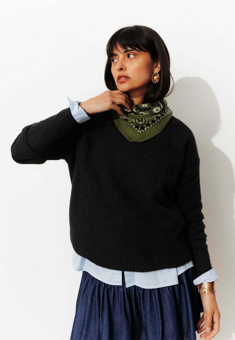 Un pull en laine noir sur une chemise à col bleu clair, associé à une écharpe à motifs verts et une jupe en jean. Des boucles d'oreilles en or ajoutent des accents.
