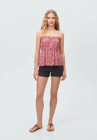 Red patterned peplum top with a smocked bodice, thin straps, paired with dark shorts and brown flip-flops on a light background.