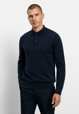 Mann mit kurzen Haaren, der ein dunkelblaues Langarm-Poloshirt und dunkle Hosen trägt, steht mit einer Hand in der Tasche vor einem einfarbigen Hintergrund.