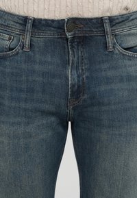 Primer plano de la cintura de un pantalón vaquero azul con botón y cremallera parcialmente bajada, usado con un suéter de punto en color crema.