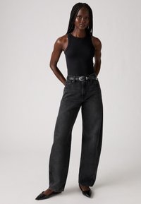 Débardeur noir à côtes associé à un jean noir à taille haute et jambes larges. Silhouette ajustée avec une ceinture noire et des escarpins noirs pointus.