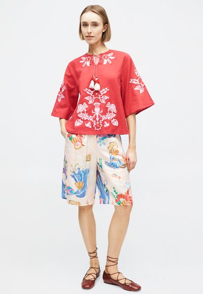 Femme portant un chemisier rouge brodé, un short coloré imprimé arrivant aux genoux, et des chaussures plates marron à lacets, debout devant un fond uni.