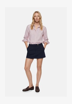 Kvinde iført en lyserød og hvid stribet langærmet skjorte stoppet ned i en navy kort nederdel med forlommer og brune loafers.