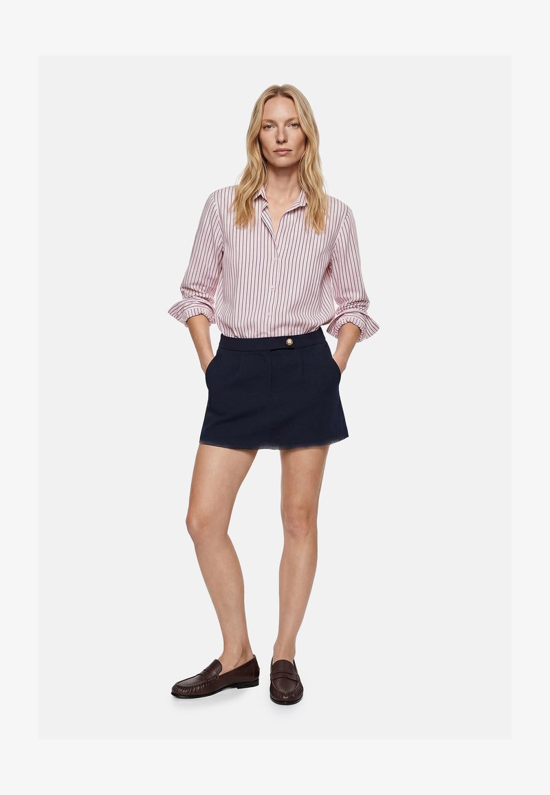 Mujer que lleva una camisa de manga larga a rayas rosas y blancas metida en una falda corta azul marino con bolsillos delanteros y mocasines marrones.