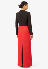Giacca nera corta con dettaglio in pizzo sul retro indossata sopra una lunga gonna rossa con uno spacco sul retro. Il design complessivo è elegante e aderente.