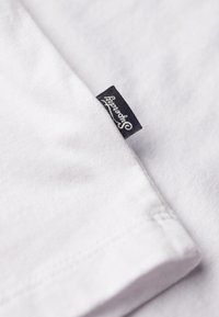 Hvid bomulds T-shirt med en glat tekstur, prydet med en marineblå label syet på venstre side, som viser mærkenavnet i hvid skrift.