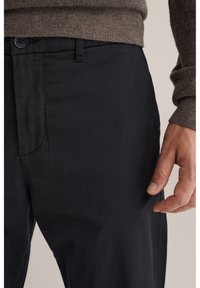 Primer plano de una persona que lleva pantalones negros y un suéter de punto marrón, con la mano derecha relajada a su lado.