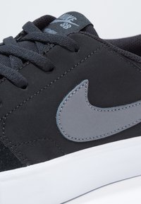 Mustat skeittikengät, joissa on mokkanahkainen yläosa, harmaa Nike-kaarre ja valkoinen kumipohja. Näkyvät silmukkareiät ja teksturoitu nauhoitus ylhäällä.