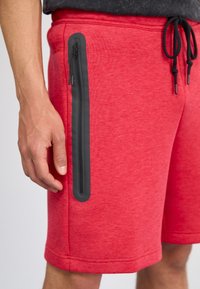 Červené sportovní šortky vyrobené z měkkého materiálu, s černou kapsou na zip na boku, viditelným šňůrkovým stahováním a prošitým lemem.