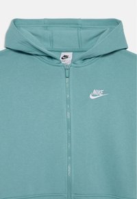 Šviesiai žydros spalvos užtraukiama džemperis, pagamintas iš minkštos medžiagos. Turi gobtuvą, balta „Nike“ logotipą išsiuvinėtą ant krūtinės ir matomas siūlų siuvimas.