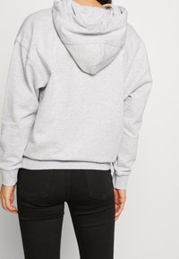 Světle šedá mikina s kapucí, volného střihu, s žebrovanými manžetami a pasem. Má jednoduchý design a měkkou texturu, kombinována s černými džíny.