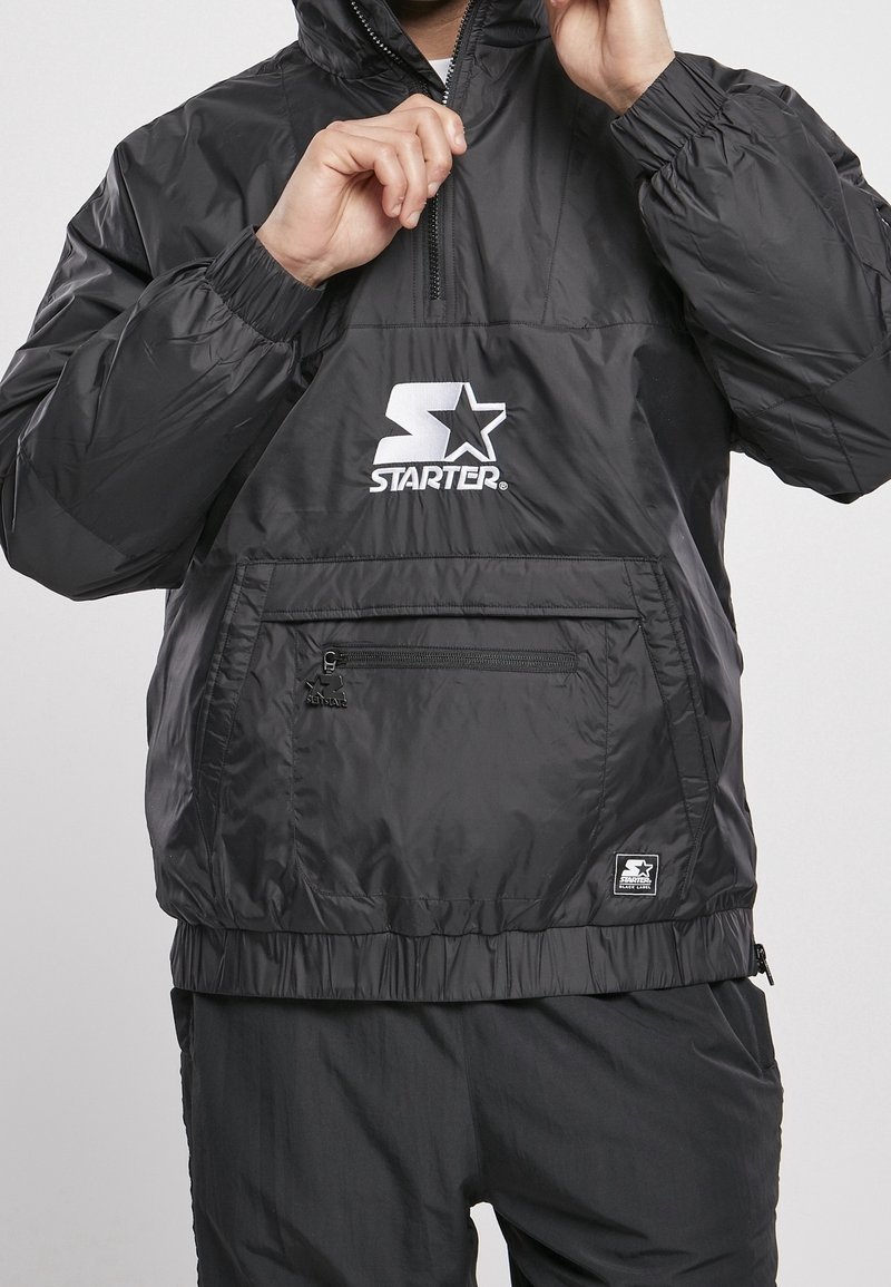 Black windbreaker jacket made of lightweight material, featuring a front pocket, elastic cuffs, and a white "Starter" logo.