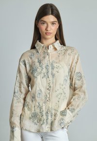 Blusa leggera color beige chiaro a fantasia floreale, con maniche lunghe, chiusura a bottoni e ricami blu intricati sul tessuto. Texture liscia e leggera.