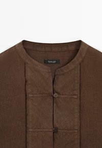Een bruine linnen shirt met een Mandarin-kraag, voorzien van een gedeeltelijke knoopsluiting en decoratieve panelen, met een gladde textuur en een eenvoudig ontwerp.