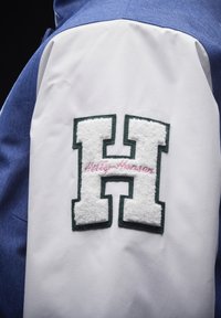 Blauw en witte jacksleeve met een grote, donzige witte "H" patch omlijnd in het groen, met "Holly Hansen" geborduurd in het roze.