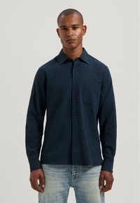 Donkerblauwe overhemd met lange mouwen, van een gestructureerde stof, met knoopsluiting, puntkraag en een borstzak. Gepaarde met lichte denim jeans.