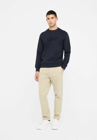 Marineblauer Sweatshirt mit geprägtem LIUJO-Logo, aus Baumwollmischmaterial, Rundhalsausschnitt, gerippte Bündchen, kombiniert mit beigen Hosen und weißen Sneakers.