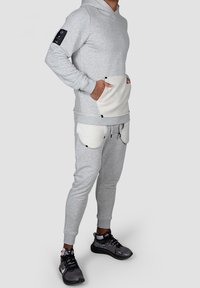 Sudadera gris con bolsillo tipo canguro y parche negro, combinada con pantalones deportivos grises que cuentan con bolsillos curvados y cordón. Textura mate.