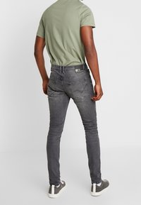 Jeans ajustados grises con una textura suave, que cuentan con dos bolsillos traseros y una etiqueta negra en la cintura, combinados con una camiseta verde y zapatillas grises.