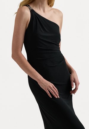 Vrouw draagt een nauwsluitende zwarte jurk met één schouder en een decoratieve kettingband aan de linkerschouder tegen een effen achtergrond.