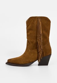 Botte de cowboy en suede marron avec un bout pointu, un talon empilé et un long détail de franges sur le côté.