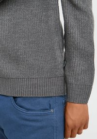 Pulover gri din tricot, cu manșete și tiv riblate, având un finisaj texturat. Asortat cu pantaloni albaștri, arătând o vedere laterală a ținutei.
