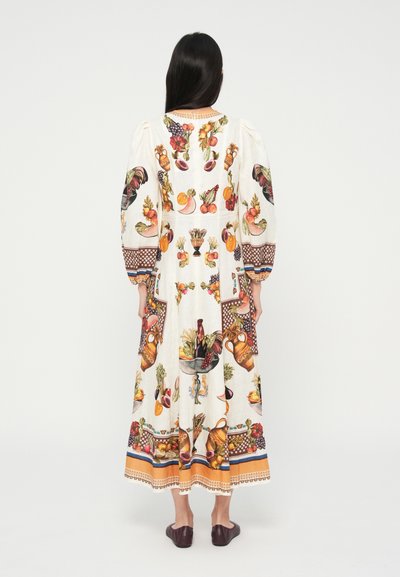 Femeie cu păr lung și închis la culoare, purtând o rochie albă lungă cu imprimeuri colorate de fructe, legume și ceramică, stând cu spatele.