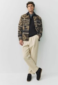 Giovane uomo appoggiato a un muro, indossa un giubbotto marrone a fantasia, un pullover nero, pantaloni beige e scarpe nere.