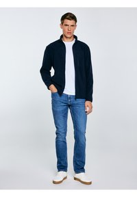 Młody mężczyzna w granatowej kurtce na zamek, białej koszulce, niebieskich dżinsach i białych trampkach, stojący z jedną ręką w kieszeni na jasnym tle.