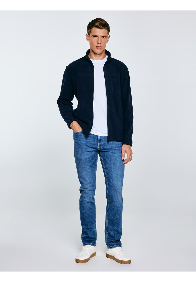 Młody mężczyzna w granatowej kurtce na zamek, białej koszulce, niebieskich dżinsach i białych trampkach, stojący z jedną ręką w kieszeni na jasnym tle.