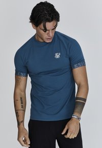 Camiseta azul de cuello redondo y mangas cortas, con un pequeño logo blanco en el pecho y detalles estampados en los puños de las mangas.