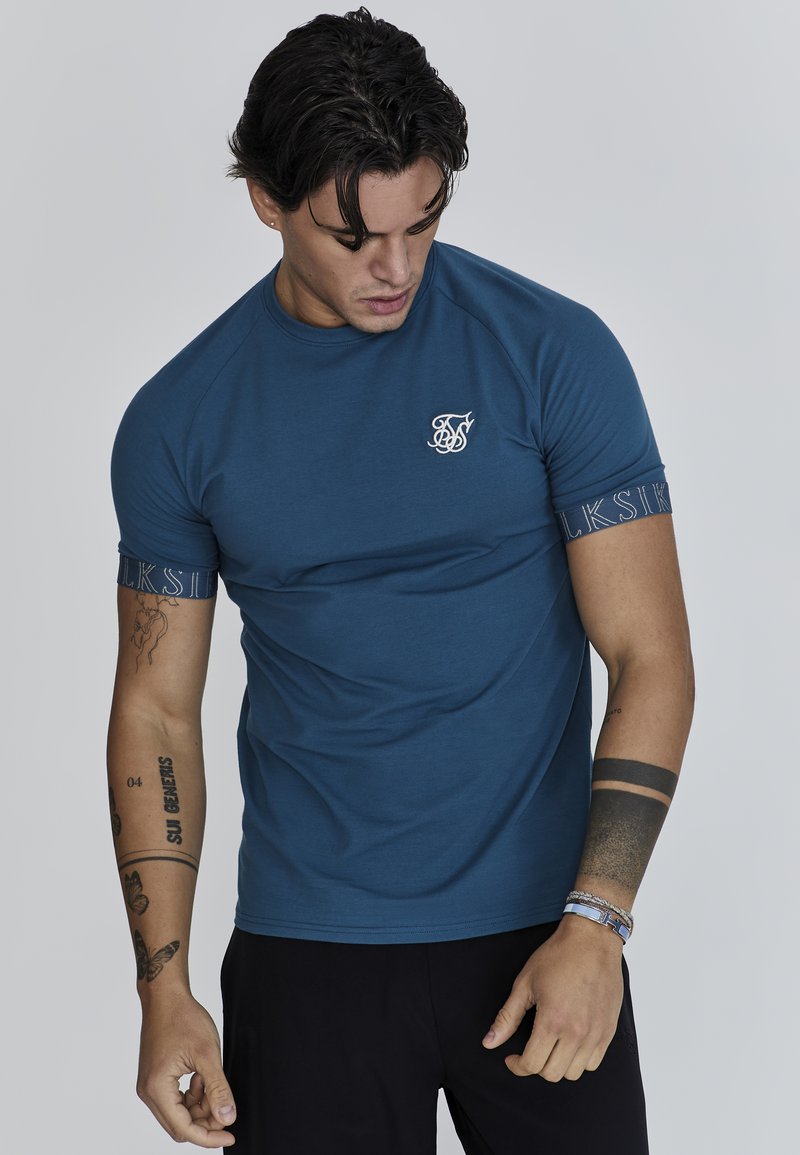 Camiseta azul de cuello redondo y mangas cortas, con un pequeño logo blanco en el pecho y detalles estampados en los puños de las mangas.