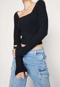 Sort ribbet croptop med firkantet halsudskæring og lange ærmer. Stoffet er tekstureret; ærmerne har tommelhul-detaljer. Båret med lyseblå jeans.