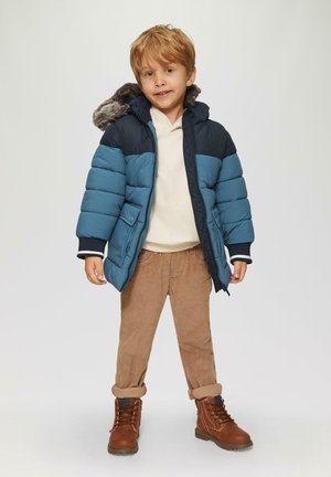 Doudoune en bleu marine et en bleu canard avec une capuche en fausse fourrure, assortie à un pantalon en velours côtelé beige et des bottes à lacets marron.