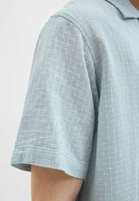 Camisa de manga corta azul claro hecha de tejido texturizado, que presenta un patrón de cuadros con sutiles costuras blancas y un cuello clásico.