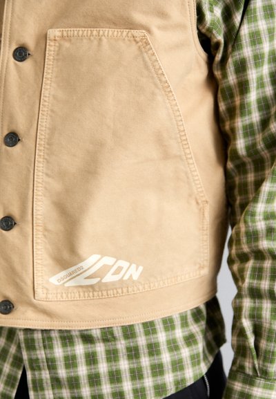 Tan canvas vest with a front pocket, button closure, and white branding. Worn over a green and white checkered shirt.