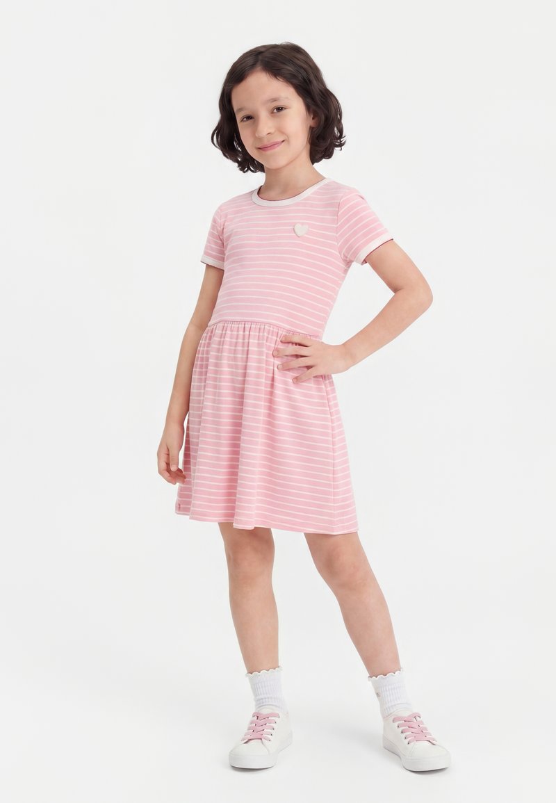 Jeune fille aux cheveux foncés mi-longs souriante, portant une robe rayée rose et blanche et des baskets blanches avec des lacets roses.