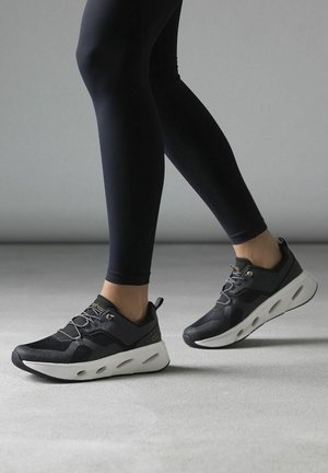 Black athletic shoes with a textured upper, grey accents, and a white, cushioned sole, paired with black leggings against a neutral background.
