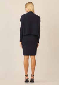 Navy-blaues geripptes Strickkleid mit einem kurz geschnittenen Bolero-ähnlichen Overlay. Es hat lange Ärmel und eine figurbetonte Silhouette, kombiniert mit schwarzen Riemchenhighheels.