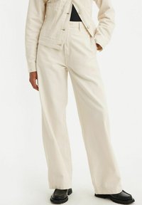 Crème wijd-uitlopende broek gemaakt van een katoenmix, met een hoge taille, zijzakken en een soepele textuur, gecombineerd met zwarte schoenen.