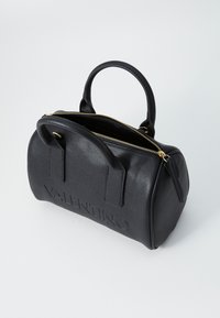 Sac à main en cuir noir avec fermeture éclair dorée, en forme arrondie, et nom de marque en relief. Deux poignées pour le porter, surface texturée.