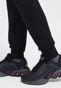 Chaussures de sport noires avec tige en mesh et accents violets, dotées d'un amorti à bulles. Associées à un pantalon de survêtement noir, mettant en valeur des poignets élastiques.
