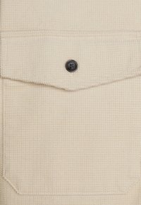 Beige textured fabric with a pocket flap, featuring a black button closure. The surface shows a small, raised pattern.