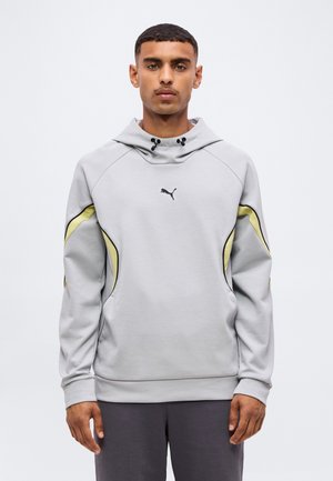 Jeune homme portant un sweat à capuche Puma gris clair avec des accents jaunes et noirs, debout sur un fond blanc uni, face à l'avant.