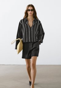 Zwarte langemouwen shirt met witte verticale strepen, gecombineerd met zwarte shorts. Houdt een rieten tas vast en draagt donkere zonnebrillen. Gladde stoftextuur.