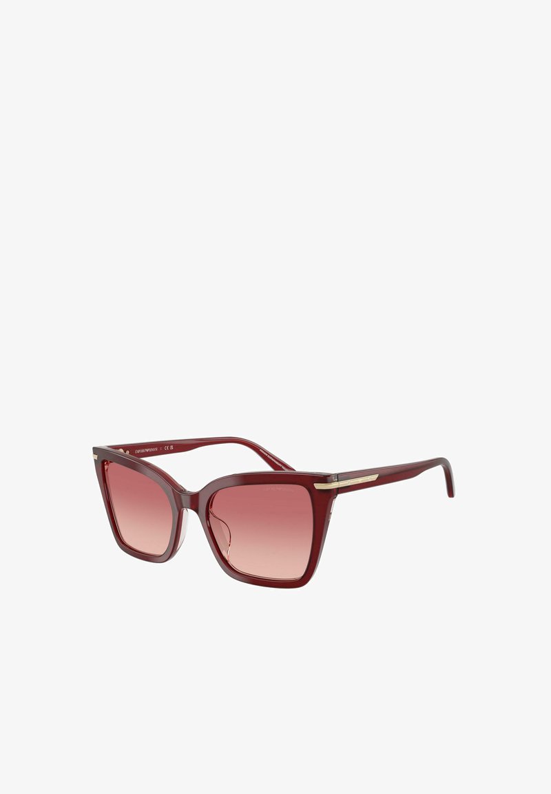 Gafas de sol rojas translúcidas de tipo ojo de gato con lentes rosas degradadas y un delgado detalle dorado en las patillas, inclinadas para mostrar detalles frontales y laterales.