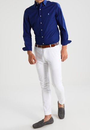 Man draagt een donkerblauw overhemd met knopen, witte slim-fit broek, bruine riem en grijze loafers, staande tegen een egale lichte achtergrond.