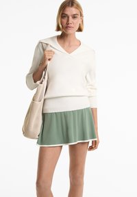Vrouw in een witte V-hals trui, groene rok met witte bies en een beige gaas schoudertas.
