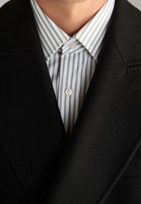 Blazer nero su misura con rever affilati indossato sopra una camicia a righe blu e bianche con bottoni. Presenta una texture liscia e una leggera lucentezza.