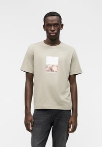 Ανεπίσημο κοντομάνικο t-shirt σε ανοιχτό ελιά, με ορθογώνιο γραφικό φύσης και λεπτομέρεια κειμένου στο κάτω μέρος. Κατασκευασμένο από απαλό βαμβακερό υλικό.